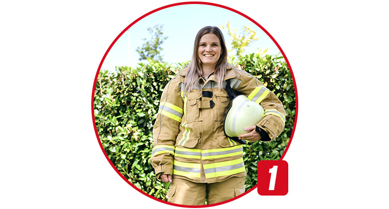 Eine junge Frau in kompletter Feuerwehrschutzkleidung hält einen Feuerwehrhelm in der Hand.