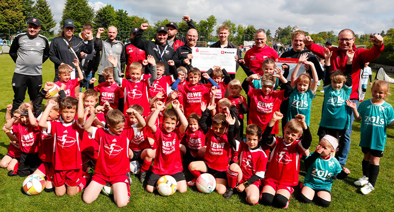 Eine große Gruppe Kinder in Fußballtrikots jubelt mit ihren Trainer:innen auf einem Fußballfeld.