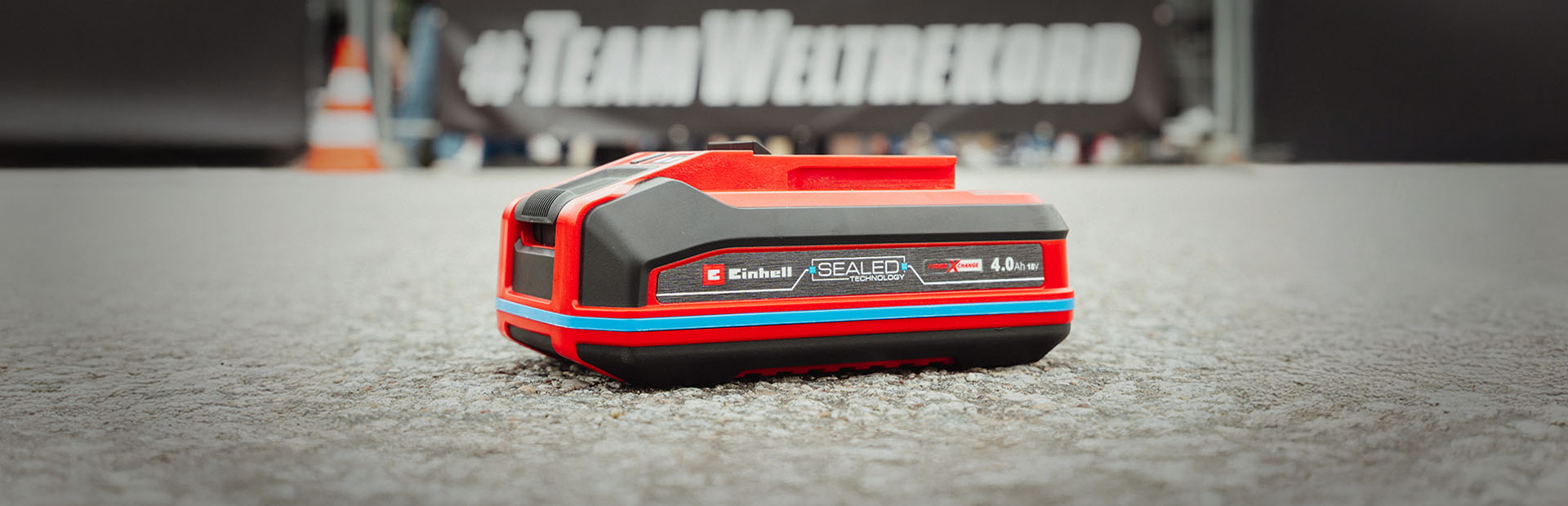 Einhell SEALED Akku Power X-Change 4,0 Ah liegt auf Asphalt. Im Hintergrund steht verschwommen "#TeamWeltrekord".