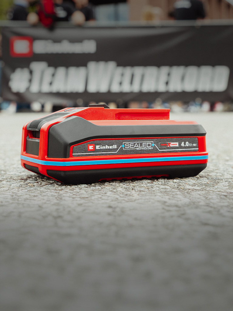 Einhell SEALED Akku Power X-Change 4,0 Ah liegt auf Asphalt. Im Hintergrund steht verschwommen "#TeamWeltrekord".