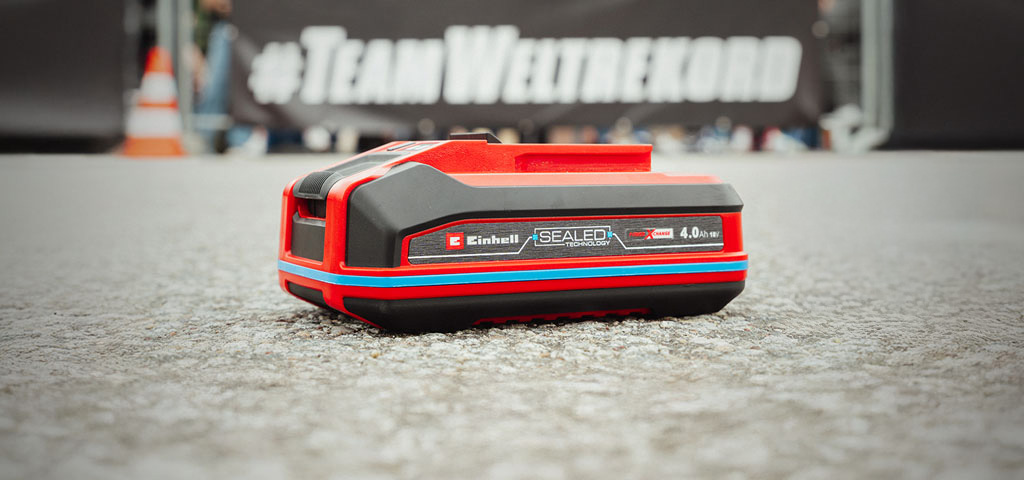 Einhell SEALED Akku Power X-Change 4,0 Ah liegt auf Asphalt. Im Hintergrund steht verschwommen "#TeamWeltrekord".