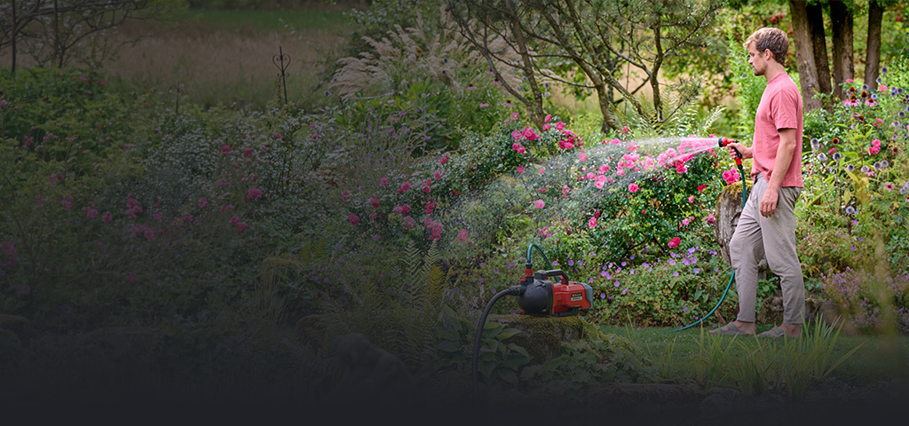 Ein Mann gießt mit einem Schlauch eine rot blühende Hecke, die an eine rote Einhell Gartenpumpe angeschlossen ist.