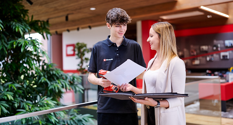 Ein junger Mitarbeiter im Einhell-Poloshirt hält einen Akkuschrauber und bespricht Dokumente mit einer Kollegin in einem modernen Büro.