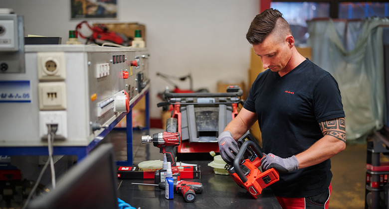 Ein Handwerker in schwarzer Einhell-Kleidung montiert oder repariert an einem Prüfstand ein rotes Einhell-Gerät.